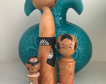 May include: Three wooden Japanese kokeshi dolls, a woman, a man, and a child, with painted faces and traditional clothing. The dolls are standing in front of a teal blue ceramic vase.