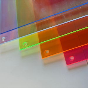 May include: Five clear plastic rulers stacked on top of each other. The rulers are different colors: blue, purple, green, orange, and pink. Each ruler has a hole in the top.