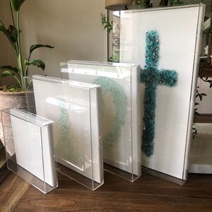 May include: Four clear acrylic frames with white mats and different sized canvases. The largest canvas features a turquoise blue cross made of glass mosaic tiles.