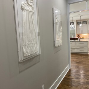 May include: Two framed white garments displayed on a hallway wall with a wooden floor. The hallway has a white trim and a light gray wall. The hallway leads to a kitchen with white cabinets and a wood floor.