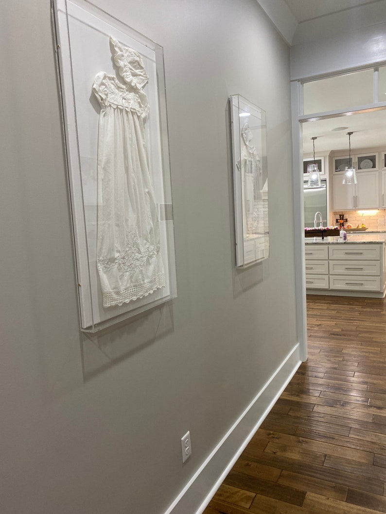 May include: Two framed white garments displayed on a hallway wall with a wooden floor. The hallway has a white trim and a light gray wall. The hallway leads to a kitchen with white cabinets and a wood floor.
