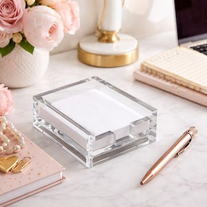 May include: A clear acrylic rectangular memo pad holder filled with white paper. A rose gold pen rests nearby, along with a pink notebook and a bouquet of pink roses in a white vase. The items are on a marble surface.