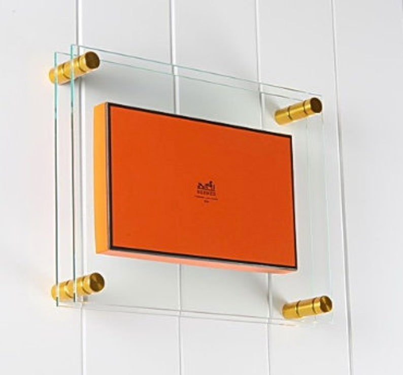 May include: A rectangular orange box with the word "Hermes" printed on it is displayed in a clear glass frame with gold metal corner brackets.