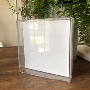 May include: A clear acrylic frame with a white canvas insert. The frame is sitting on a wooden surface.