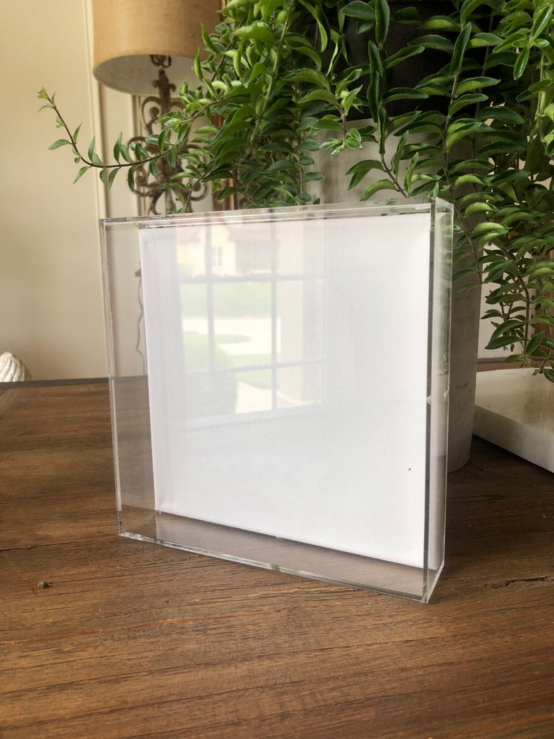 May include: A clear acrylic frame with a white canvas insert. The frame is sitting on a wooden surface.