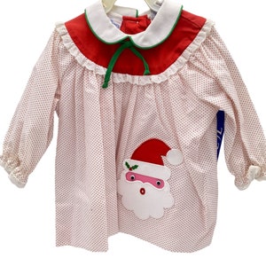 May include: A white dress with red polka dots and a red and green Santa Claus appliqué. The dress has a white collar with a green bow and a red trim.