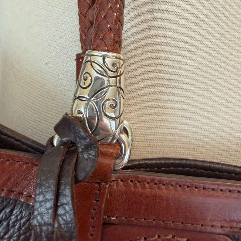 May include: Close-up of a brown leather handbag with a braided handle. The handle is attached to the bag with a decorative silver metal clasp. The bag has a dark brown and reddish-brown color scheme.
