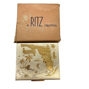 May include: A gold compact with a map of Florida and the words "A Ritz Creation" on the top. The compact is open and the inside is silver.