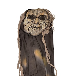 May include: A Halloween mask with a weathered, golden-brown face and dark eye sockets. The mask is attached to a dark brown fabric hood with tattered, rope-like accents. The overall design suggests a spooky, aged appearance.