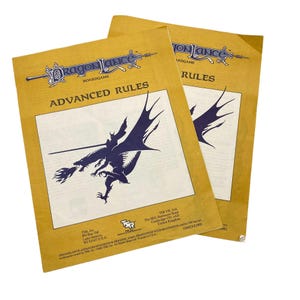 May include: Two copies of the Dragonlance Boardgame Advanced Rules book. The covers are tan with a blue and white illustration of a dragon and knight. The text "ADVANCED RULES" is visible.