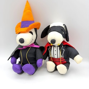 May include: Two plush Snoopy figures dressed in Halloween costumes. One wears a witch costume with an orange and purple hat, black cape, and purple accents. The other is in a tuxedo with a red cape and a black mask. Both have white bodies and black ears.