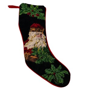 May include: Black Christmas stocking with a red trim and loop. The stocking features a needlepoint design of Santa Claus with a white beard and red hat, surrounded by green foliage and red berries.