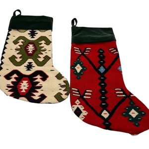 May include: Two Christmas stockings with a woven design. One stocking is off-white with green, black, and red geometric patterns. The other is red with black, blue, and white geometric patterns. Both have a dark green cuff and a loop for hanging.