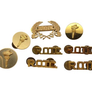 May include: A collection of gold-colored ROTC pins and badges. The pins feature the word "ROTC" and a torch design. The badges feature a wreath design with the word "ROTC" inside.