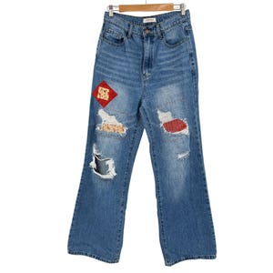 May include: Light blue denim jeans with distressed details and decorative patches. The jeans feature ripped areas and patches in red and floral patterns. The jeans are hanging on a wooden hanger.