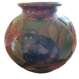 May include: A handcrafted ceramic vase with a dark blue monkey design, surrounded by leaves in shades of green, orange, and brown. The vase has a textured background and a dark red rim. This decorative vase is a unique home decor item.