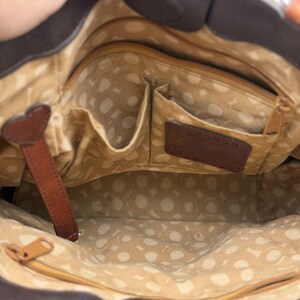 May include: Interior view of a brown leather handbag with multiple pockets and a patterned lining. The bag features a leather strap with a heart-shaped detail and a brand label. The interior is tan with white polka dots.
