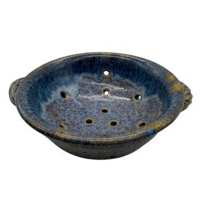 May include: A round ceramic colander featuring a mottled blue and brown glaze with a glossy finish. The bowl has two side handles and multiple drainage holes distributed across the interior base and sides, showcasing a speckled texture throughout.