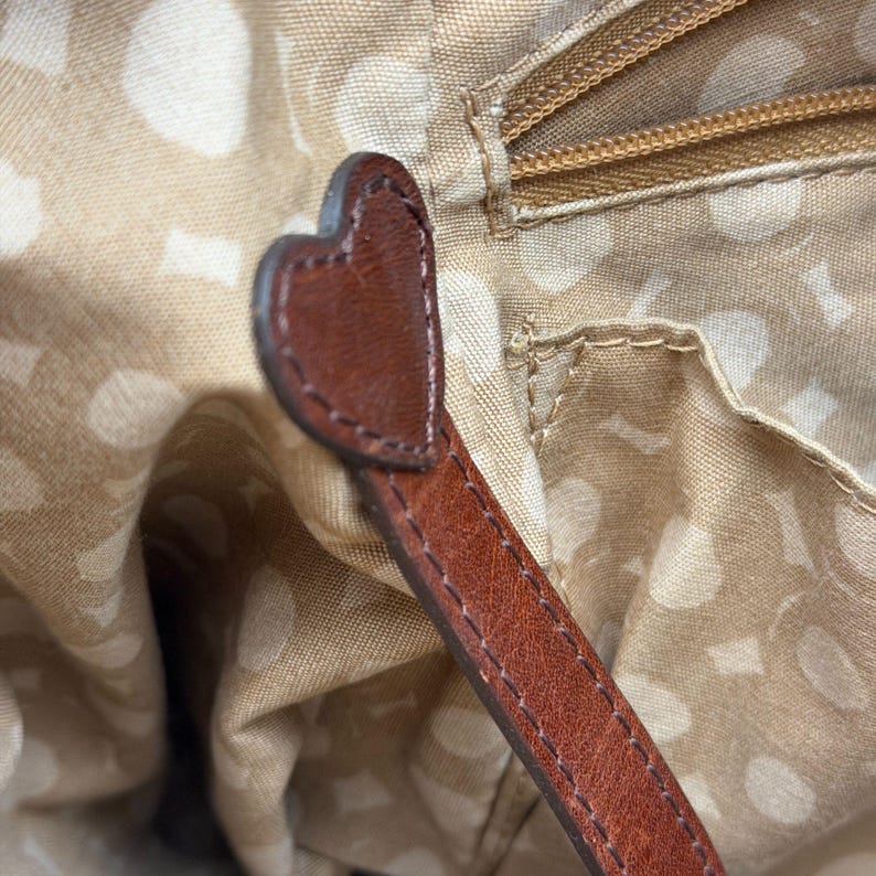 May include: Close-up of a brown leather handbag strap with a heart-shaped detail. The strap is stitched with a contrasting thread. The interior of the bag is visible, featuring a beige fabric with a white polka dot pattern.