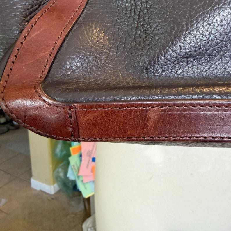 May include: Close-up of a brown leather bag with a contrasting dark brown leather trim. The bag features detailed stitching along the edges, showcasing the craftsmanship. The leather has a textured appearance.