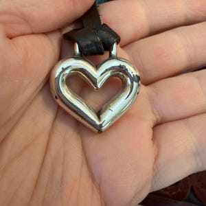May include: A silver heart-shaped pendant with a black leather cord. The pendant has a hollow center and shows signs of wear. The leather cord is looped through the top of the heart.