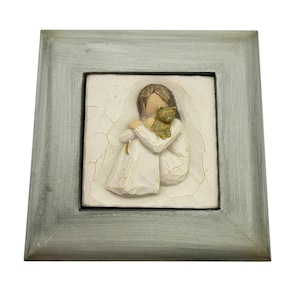 May include: A square wall plaque featuring a relief sculpture of a figure with brown hair wearing a white dress, holding a small brown cat. The sculpture is set against a cream-colored background with a crackled texture, encased in a gray wooden frame.