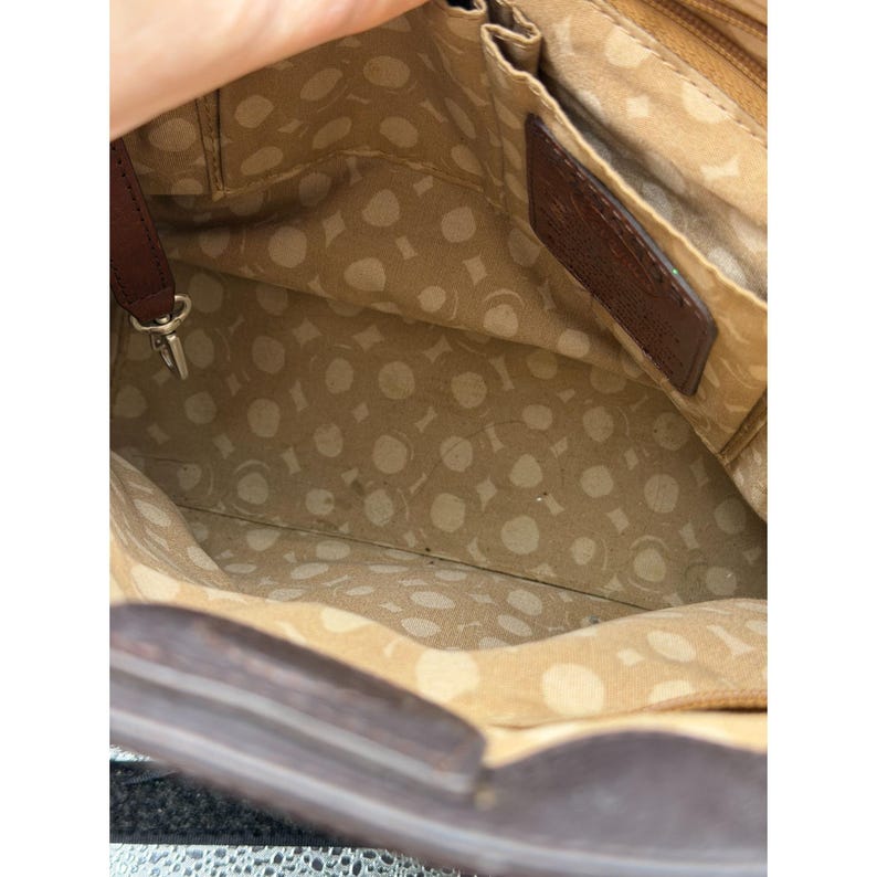 May include: Open view of a brown leather handbag's interior. The bag features a patterned tan lining with multiple compartments. A dark brown leather tag is visible. The bag's exterior is a darker shade of brown.