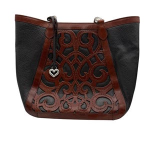 May include: Black and brown leather tote bag with a decorative cut-out design. The bag features a heart-shaped charm and brown leather trim. The tote has a structured shape and a top handle.