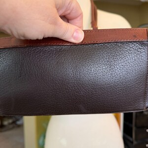 May include: A dark brown leather handbag with a textured surface and a contrasting brown leather trim. The bag is held by a hand, showcasing the top edge and handle.