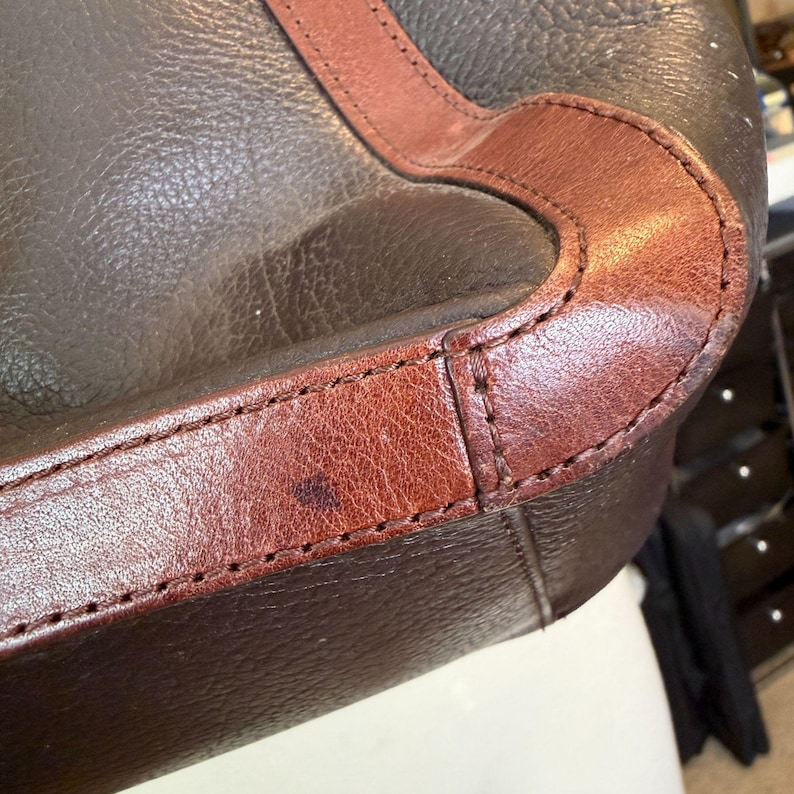 May include: Close-up of a brown leather bag with a contrasting reddish-brown trim. The bag features visible stitching and a textured surface. The leather appears worn, with a dark spot on the reddish-brown trim.
