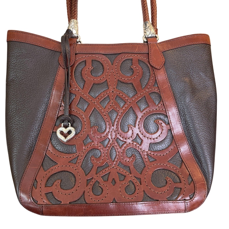 May include: A brown leather tote bag with a decorative cut-out design on the front. The bag features a dark brown body with lighter brown accents and a braided handle. A silver heart charm hangs from the handle.