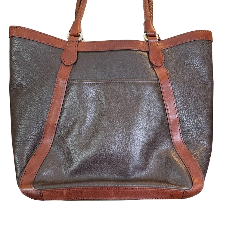 May include: A brown leather tote bag with tan leather trim and braided handles. The bag features a large main compartment and a back pocket. The leather has a textured appearance.