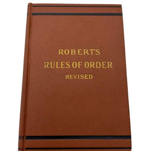 May include: A brown hardcover book titled "ROBERT'S RULES OF ORDER REVISED" in gold lettering. The book has black horizontal lines near the top and bottom edges. The book is set against a white background.