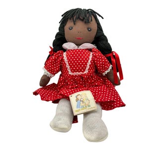May include: A handmade rag doll with dark skin, black yarn hair, and a red polka dot dress. The doll has white socks and a tag with a picture of two children. A charming and nostalgic toy.