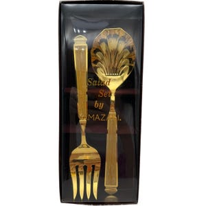 May include: A gold-toned salad set by YAMAZAKI in a rectangular box. The set includes a fork with a long handle and a spoon with a shell-shaped bowl. The text Salad Set by YAMAZAKI is printed in gold on the clear plastic packaging cover.