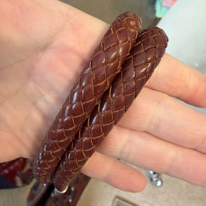 May include: Close-up of two braided brown leather straps. The straps are tightly woven, showcasing a detailed pattern. The leather has a rich, warm tone, suggesting a handcrafted item. The straps appear to be part of a larger accessory, possibly a bracelet or a bag handle.