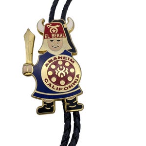 以下が含まれることがあります： 金色のボーラタイ。フェズと剣を持った漫画のようなキャラクターが特徴です。「アナハイムカリフォルニア」と「エルベカル」の文字が入った青い衣装を着ています。タイは黒い編み込みコードが付いています。
