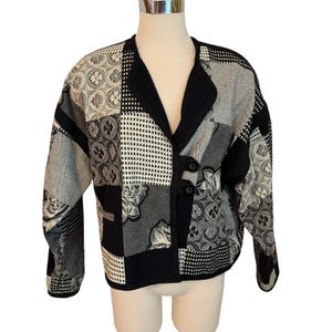May include: A long-sleeved jacket featuring a patchwork design in black, white, and gray. The garment has a V-neckline, a solid black trim, and two large black buttons on the front. The fabric displays various patterns including floral motifs, checkered squares, and herringbone textures.