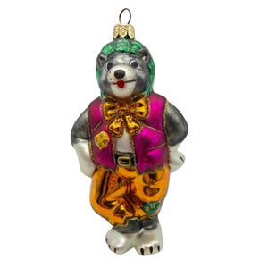 May include: A glass Christmas ornament shaped like a bear wearing a green glittery hat, a shiny magenta vest with a gold bow tie, and metallic orange trousers. The bear has a grey face, white paws, and a small gold hanging loop at the top.