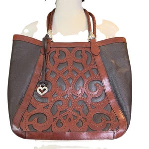 May include: A brown and tan leather tote bag with a decorative cut-out design on the front. The bag has braided handles and silver-toned hardware. A heart-shaped charm hangs from the front.
