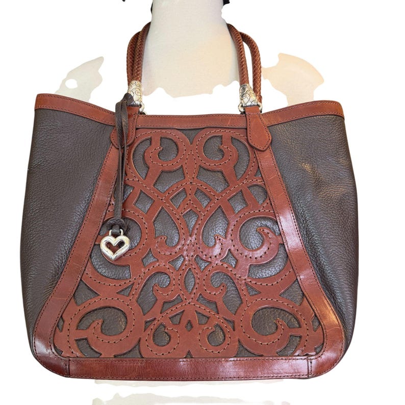 May include: A brown and tan leather tote bag with a decorative cut-out design on the front. The bag has braided handles and silver-toned hardware. A heart-shaped charm hangs from the front.