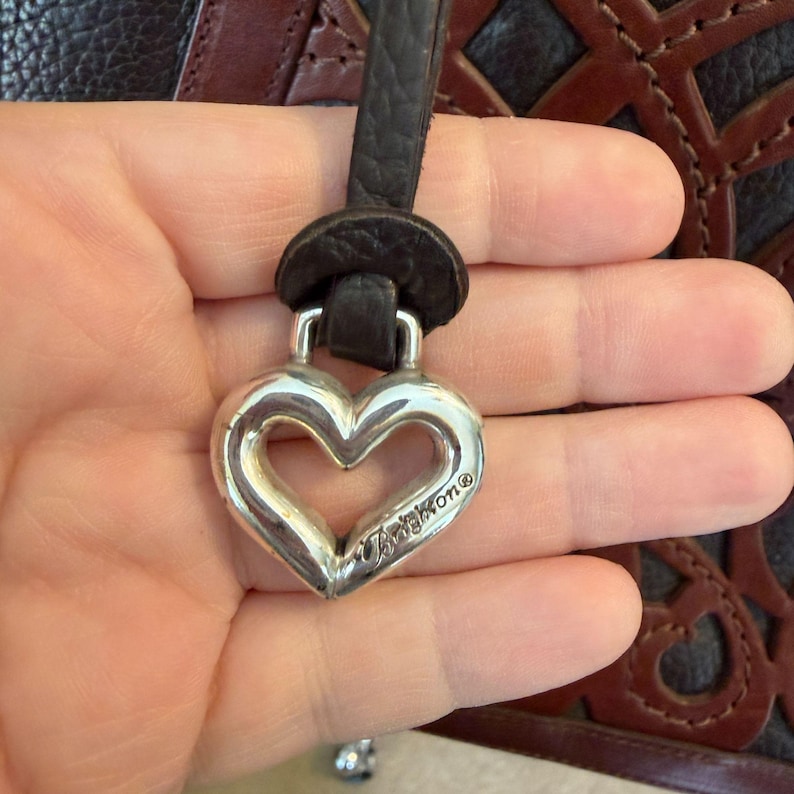 May include: Silver heart-shaped charm with the Brighton brand name, attached to a dark brown leather strap. The charm is held in front of a dark brown and black patterned background.