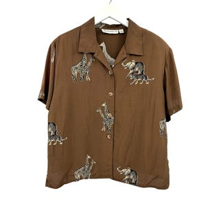 Op de afbeelding: Een bruin overhemd met korte mouwen en een safari-dierenprint. De print bevat giraffen, olifanten en tijgers. Het overhemd heeft een kraag en hangt aan een witte hanger.