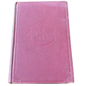 May include: A vintage, hardcover book with a textured, pink cover. The cover features a circular embossed design with a crest and decorative corner details. The book's edges show signs of wear, suggesting its age.