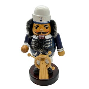 May include: A wooden sailor nutcracker figurine. The figurine wears a white hat with an anchor emblem, a navy blue jacket, and a black beard. It holds a wooden ship's wheel and stands on a dark brown base.
