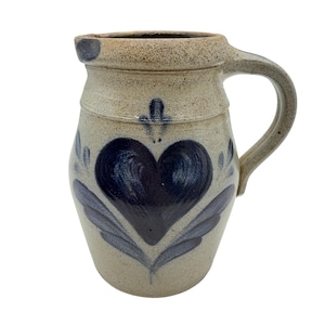 May include: A stoneware pitcher with a speckled beige finish and a single handle. The front features a hand-painted dark blue heart design with stylized leaf accents and a small floral motif above the heart, all set against a textured surface.