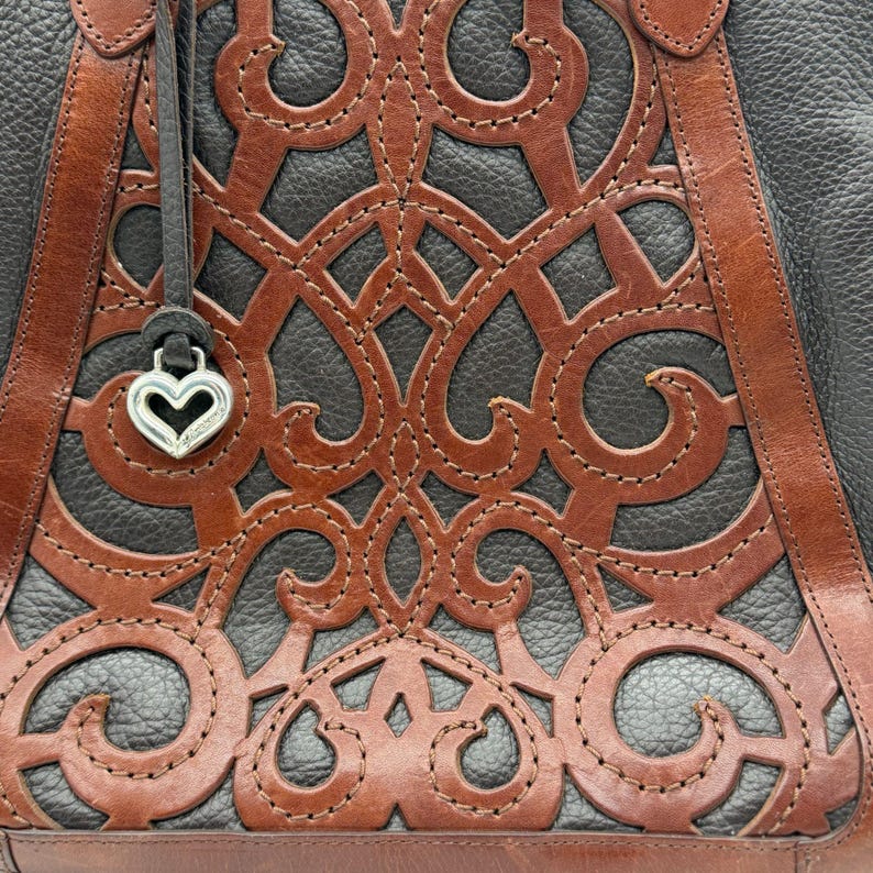 May include: Close-up of a black and brown leather handbag. The bag features a decorative cut-out design in brown leather, stitched onto a black leather base. The bag has brown leather straps and a silver heart-shaped charm.