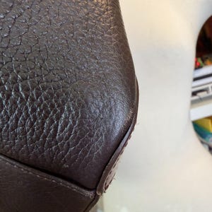 May include: Close-up of a dark brown leather bag. The leather has a textured, pebbled appearance. The bag's corner is visible, showing the stitching and construction details. The overall aesthetic suggests a durable, high-quality accessory.