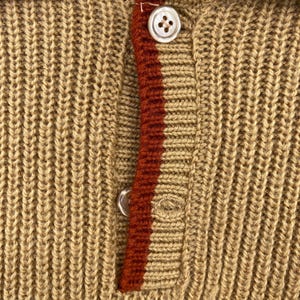 May include: A close-up of a brown knitted sweater with a red and white stripe down the front. The sweater has a button closure at the top.