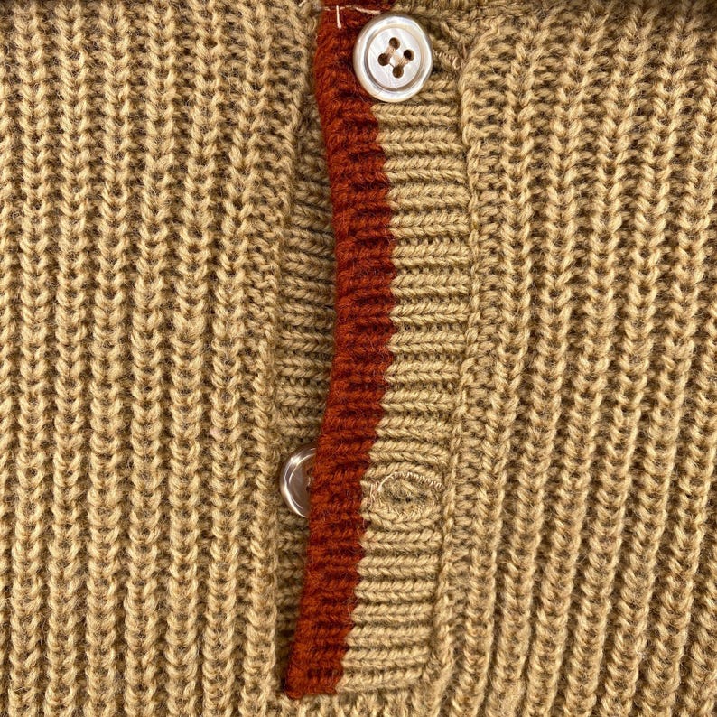 May include: A close-up of a brown knitted sweater with a red and white stripe down the front. The sweater has a button closure at the top.